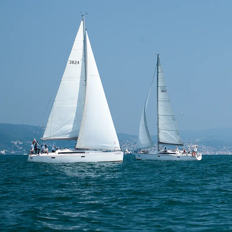 Due barche a vela in mare con persone a bordo; sullo sfondo la costa con un centro abitato