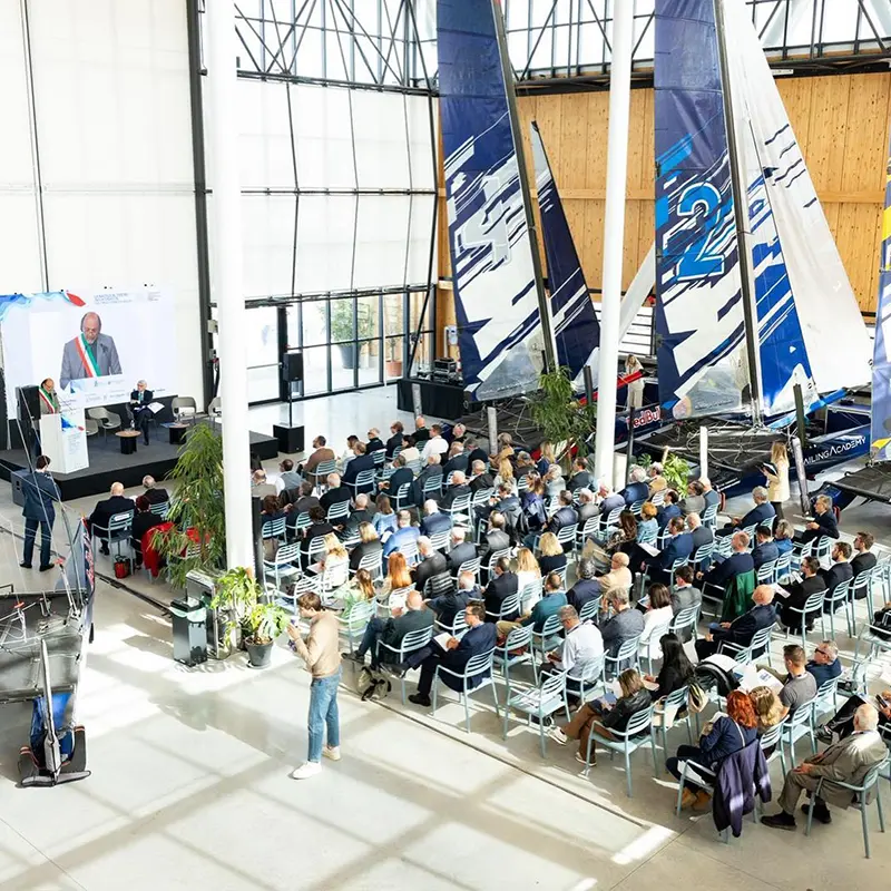 Convegno nell'hangar: pubblico seduto davanti al palco, accanto a barche a vela da regata esposte